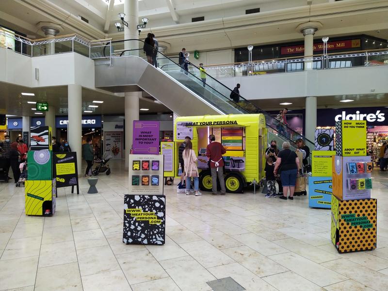 A bright yellow horsebox is set against an escalator in a shopping centre. Large colourful display boxes create a space in front of the horsebox. People are milling about it.