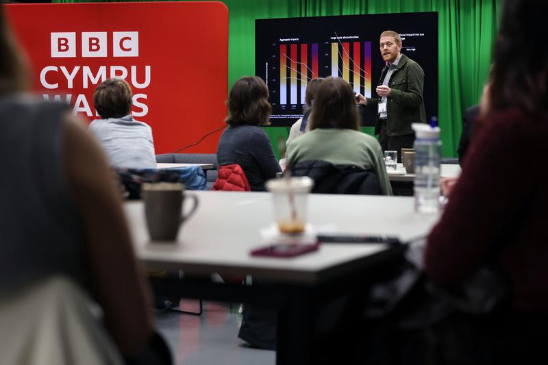 A tall white man with red hair is giving a lecture. There is a slide with graphs in the background and a large red sign reads BBC Cymru. People are sitting at desks and looking forward, towards the speaker.