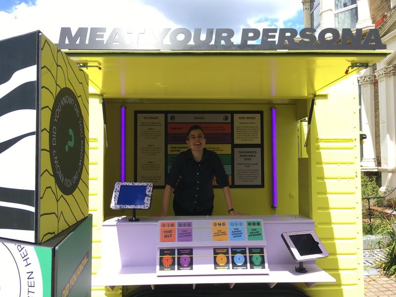 A bright yellow horsebox centres the picture with 'Meat Your Persona' written across the top. Inside a white woman with short dark hair is smiling. She stands behind lilac shelves which hold postcards. There are two tablets at either end of the horsebox.