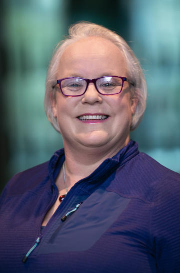 A white woman, wearing a dark blue shirt, smiles at the camera. She has glasses and grey-ish short hair.