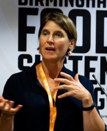 Anna taylor A white woman with short dark hair is caught talking on camera. Her mouth is slightly open, as if in the middle of saying something, and her hands are expressive in front of her. She wears a black shirt and has a gold lanyard around her neck.