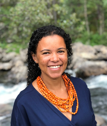 A woman with dark curly hair smiles at the camera. She wears a coral necklace and dark blue skirt.