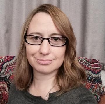 A white women smiles at the camera. She has long brown hair that reaches her shoulders and is wearing dark-rimmed glasses.