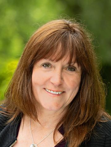 A white woman with shoulder-length brown hair and a fringe smiles at the camera. There is a silver necklace around her neck. 