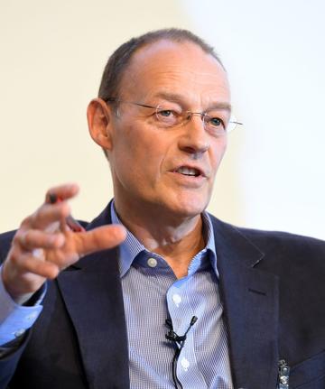 Martin white A white man is looking beyond the camera to an audience, his mouth open in speech. He has very short dark hair and wears glasses. He is dressed in a blue shirt and darker blue blazer.