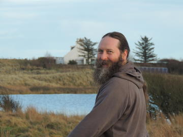 Pete smith A white man with long hair tied back, looks back towards the camera from over his shoulder. He is in a rural location with a loch and grasses in the background.