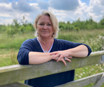 Sue pritchard headshot A white woman leans over a farm gate. She has bobbed blonde hair and wears a dark blue v-neck jersey. Behind her is a green field and blue sky with clouds.