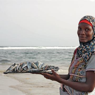 A dark skinned woman stands looking at the camera, the sea behind. She holds a flat plate of fish.