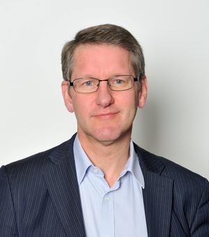 A white man with blonde hair and blue eyes smiles at the camera. He wears glasses and a blue shirt and striped blazer.