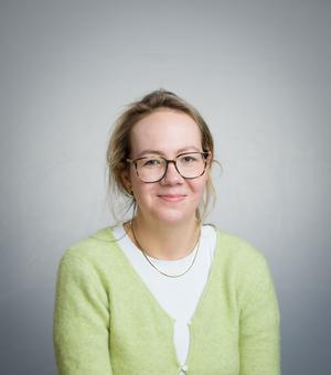 A white woman with blonde hair smiles at the camera. She is wearing a yellow cardigan and glasses.
