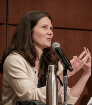 A white woman with dark hair and wearing a cream cardigan, talks with her hands. She has a microphone in front of her.