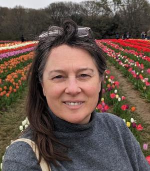 A white woman with dark hair and a grey poloneck jumper on stands in a field of tulips. She has glasses perched on her head and is smiling at the camera.