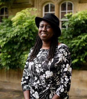 A black woman with long braids smiles at the camera. She wears a black hat and a black and white patterned dress.