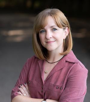A red haired white woman smiles at the camera. She is wearing a magenta top and has her arms crossed.