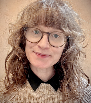 A white woman with dark curly hair and blue eyes smiles toward the camera. She is wearing glasses and a beige fisherman jersey.