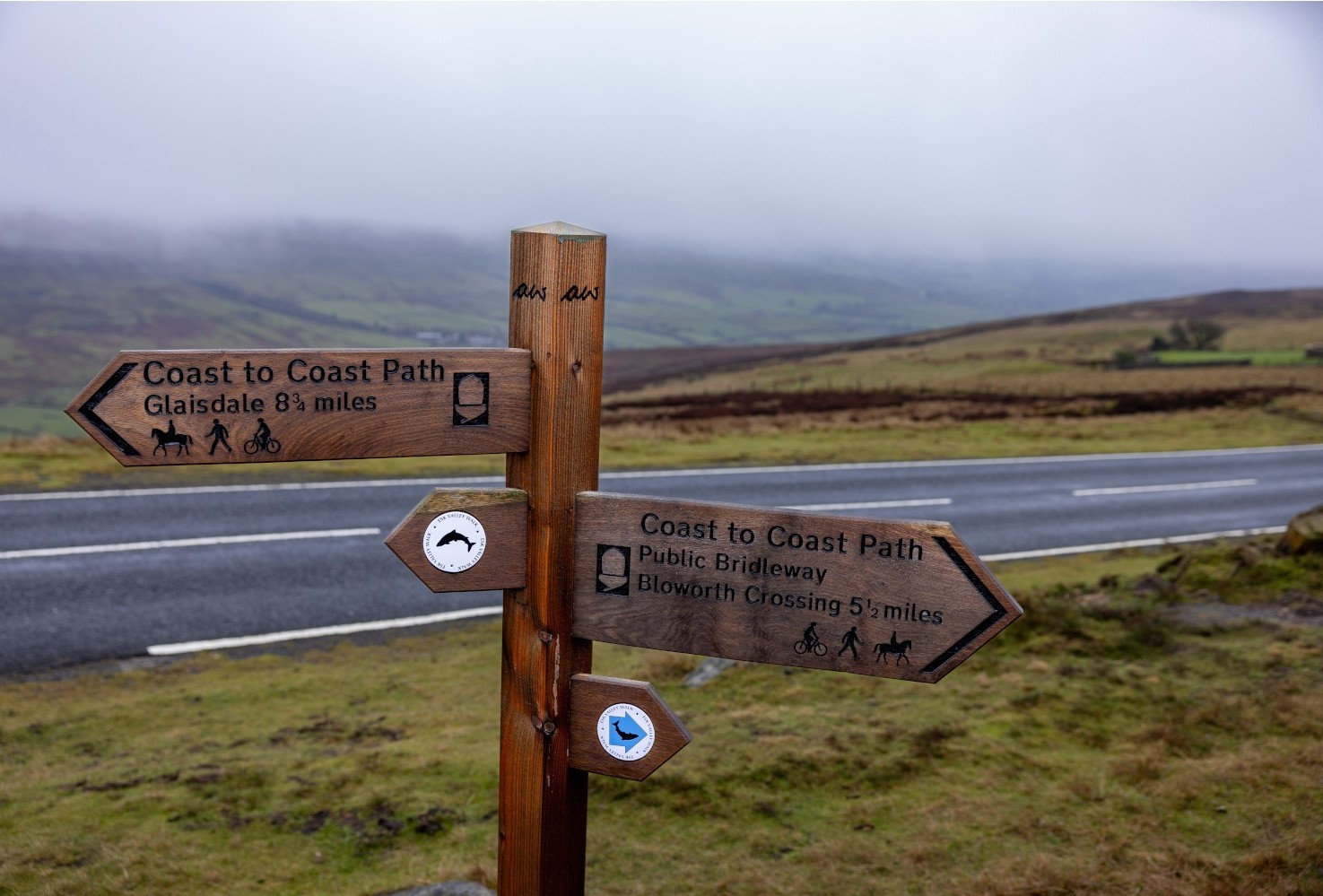 A sign showing the directions for the Wainwright Way, including the distinctive 'AW' initials at the top of the sign. Behind the sign is road and then rolling hills shrouded in mist.