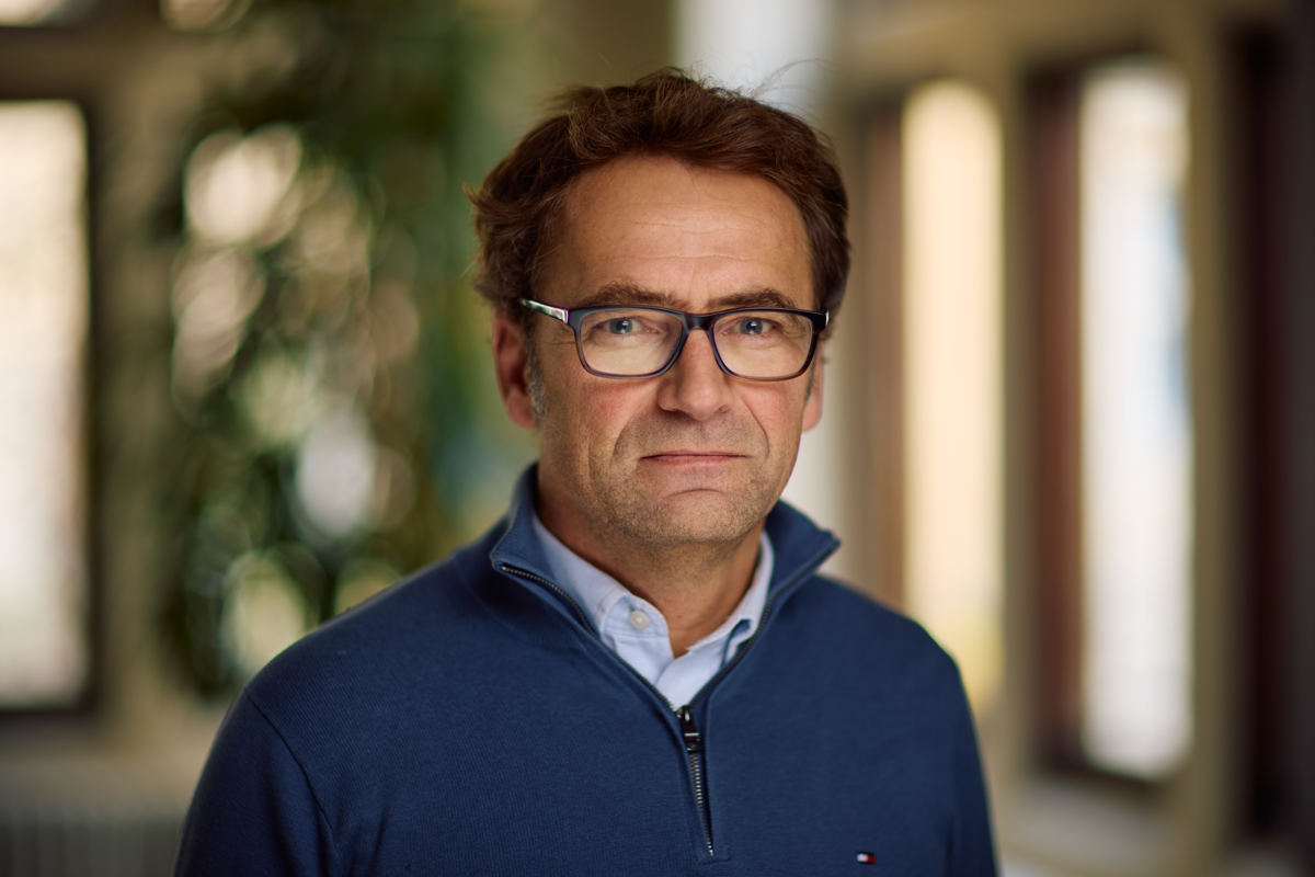 A white man with dark brown hair stares at the camera. He wears glasses and has a navy jumper over a blue shirt.