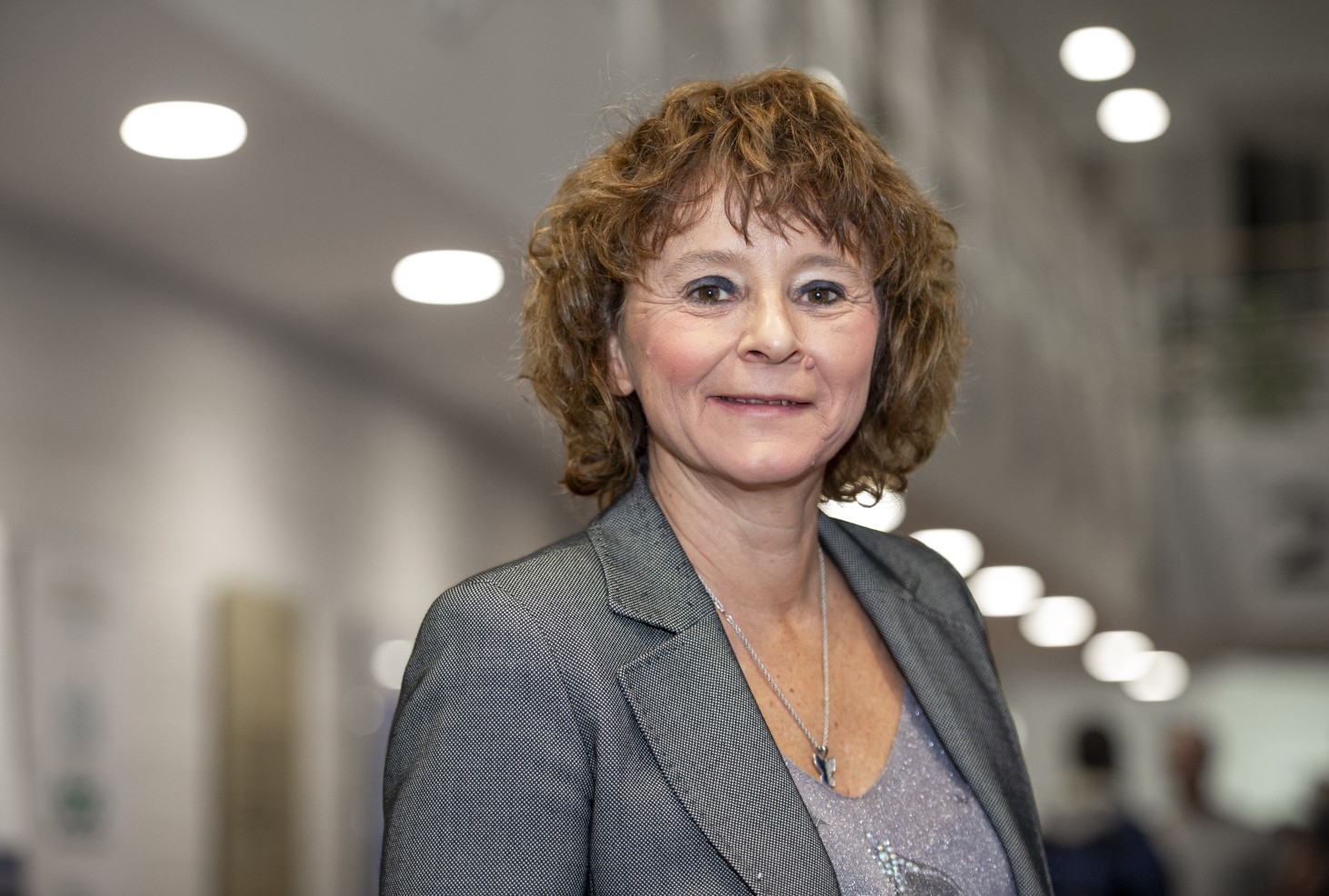 A white woman smiles at the camera. She has curly brown hair and wears a grey blazer.