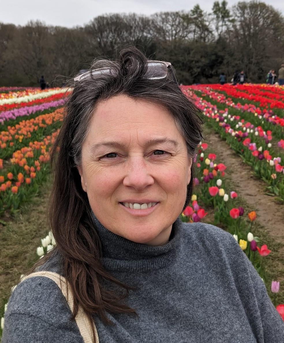 A white woman with dark hair and a grey poloneck jumper on stands in a field of tulips. She has glasses perched on her head and is smiling at the camera.