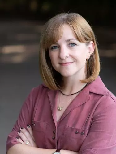 A red haired white woman smiles at the camera. She is wearing a magenta top and has her arms crossed.