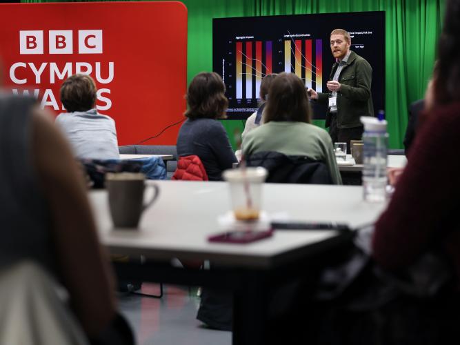 A tall white man with red hair is giving a lecture. There is a slide with graphs in the background and a large red sign reads BBC Cymru. People are sitting at desks and looking forward, towards the speaker.