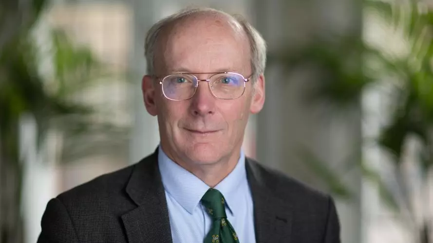A white man wearing glasses smiles at the camera. He is dressed in a suit with a green tie.
