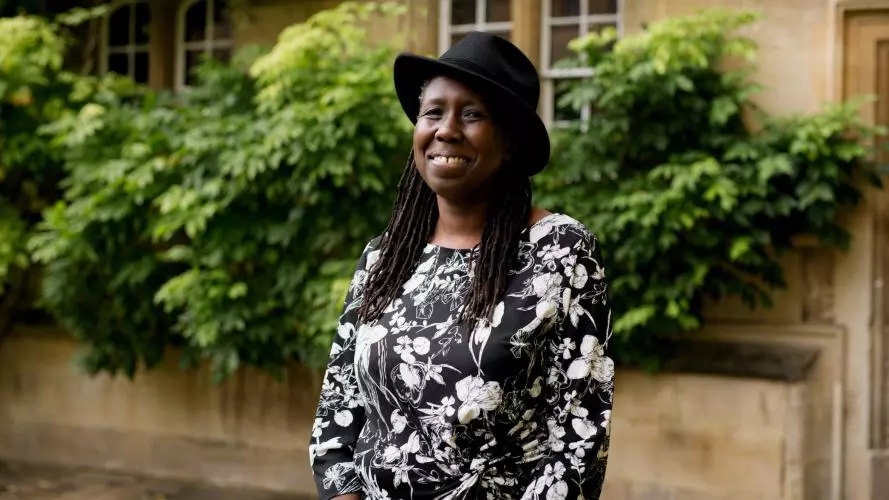 A black woman with long braids smiles at the camera. She wears a black hat and a black and white patterned dress.