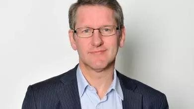 A white man with blonde hair and blue eyes smiles at the camera. He wears glasses and a blue shirt and striped blazer.