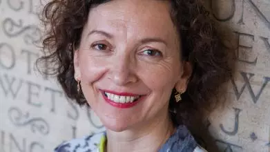 A white woman with brown curly hair smiles at the camera. She wears a blue and yellow-green shirt.