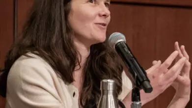 A white woman with dark hair and wearing a cream cardigan, talks with her hands. She has a microphone in front of her.