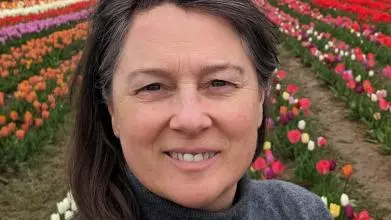 A white woman with dark hair and a grey poloneck jumper on stands in a field of tulips. She has glasses perched on her head and is smiling at the camera.