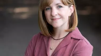 A red haired white woman smiles at the camera. She is wearing a magenta top and has her arms crossed.