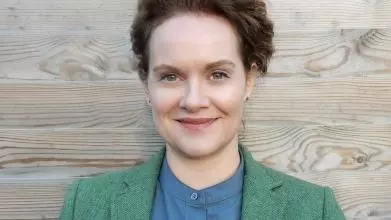 A dark-haired white woman smiles against a neutral backdrop. She is wearing a green jacket and blue shirt.