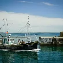A small boat enters a walled harbour. The sea is blue with some white cloud on the horizon. A lighthouse stands on the harbour wall.