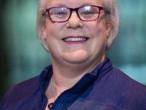 A white woman, wearing a dark blue shirt, smiles at the camera. She has glasses and grey-ish short hair.