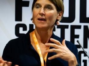 A white woman with short dark hair is caught talking on camera. Her mouth is slightly open, as if in the middle of saying something, and her hands are expressive in front of her. She wears a black shirt and has a gold lanyard around her neck.