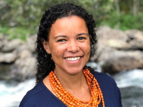 A woman with dark curly hair smiles at the camera. She wears a coral necklace and dark blue skirt.