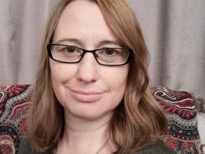 A white women smiles at the camera. She has long brown hair that reaches her shoulders and is wearing dark-rimmed glasses.