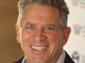A white man with short grey curly hair and blue eyes smiles at the camera. The top of his shirt collar (blue check) and blazer are visible.