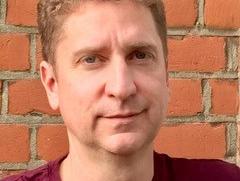 A white man with short curly blonde hair smiles at the camera. He is wearing a maroon t-shirt.