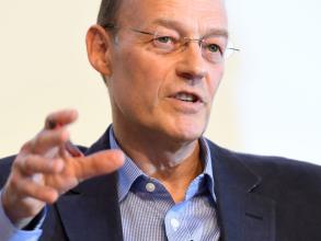 A white man is looking beyond the camera to an audience, his mouth open in speech. He has very short dark hair and wears glasses. He is dressed in a blue shirt and darker blue blazer. 