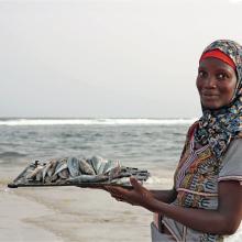 A dark skinned woman stands looking at the camera, the sea behind. She holds a flat plate of fish.