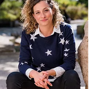A white woman with curly brown hair smiles at the camera. She is wearing a dark blue jersey with white stars, and a white collared shirt underneath.